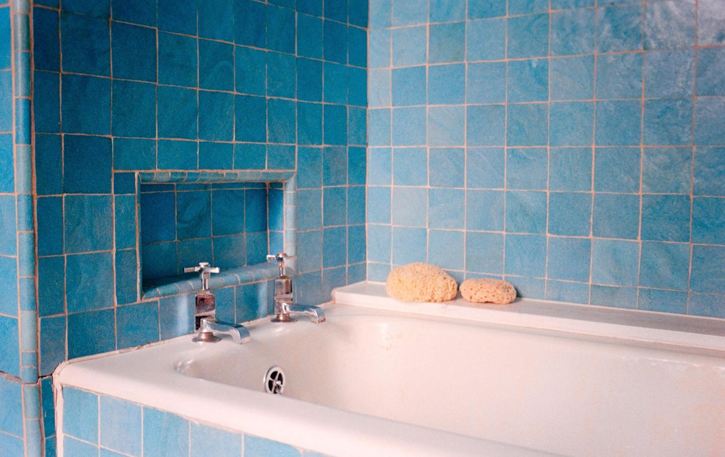 A white bathtub is surrounded by blue tiles and two sponges rest on the edge of the tub.