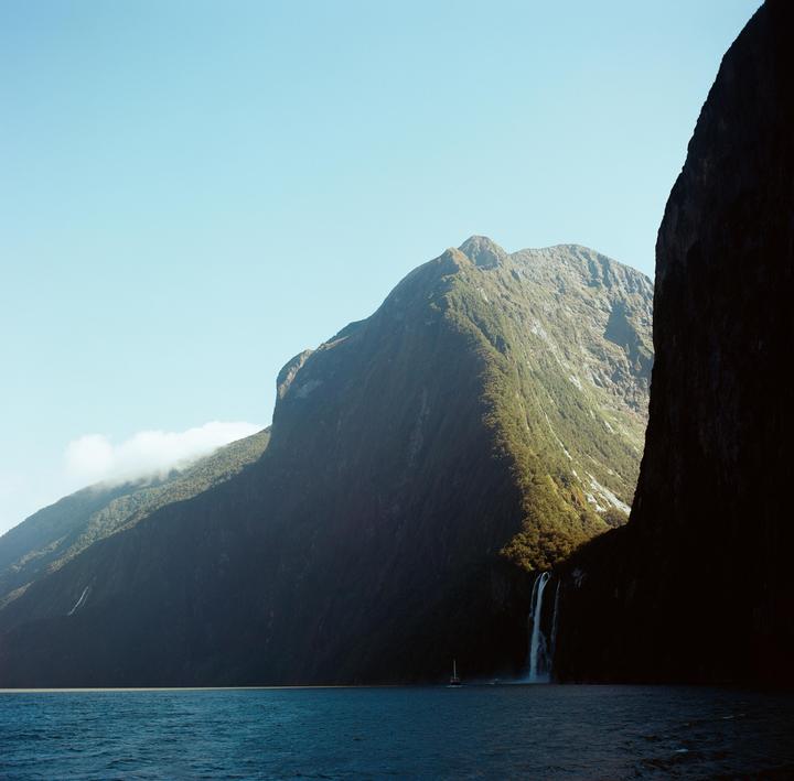 A waterfall cascades down a large mountain into a body of water with a small boat.