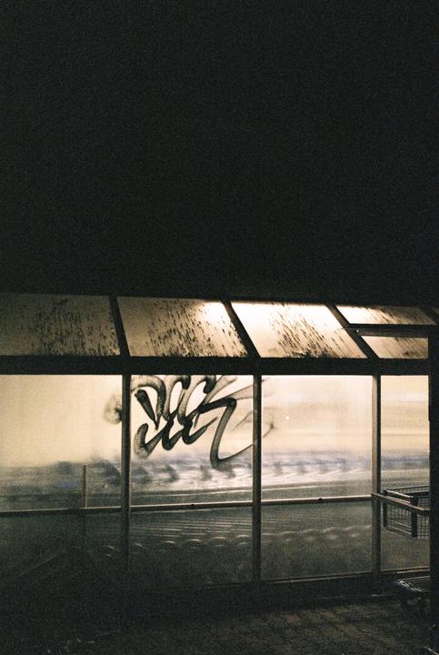 A glass shelter with graffiti on its window stands under a dark sky at night.