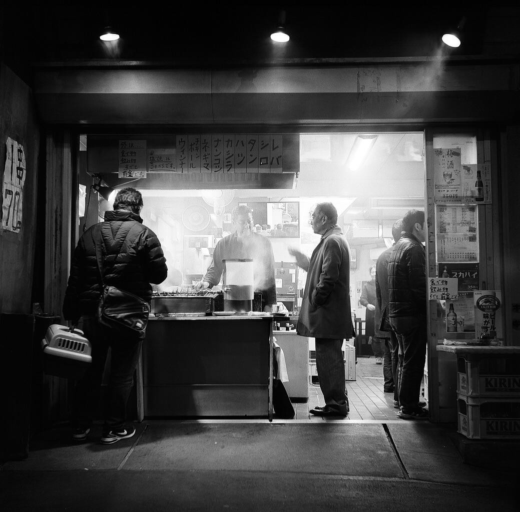 Three men stand outside a small shop at night, with one man holding a pet carrier.