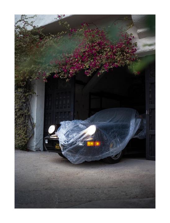 A black car covered with a plastic sheet is parked in a garage with bougainvillea growing over the entrance.