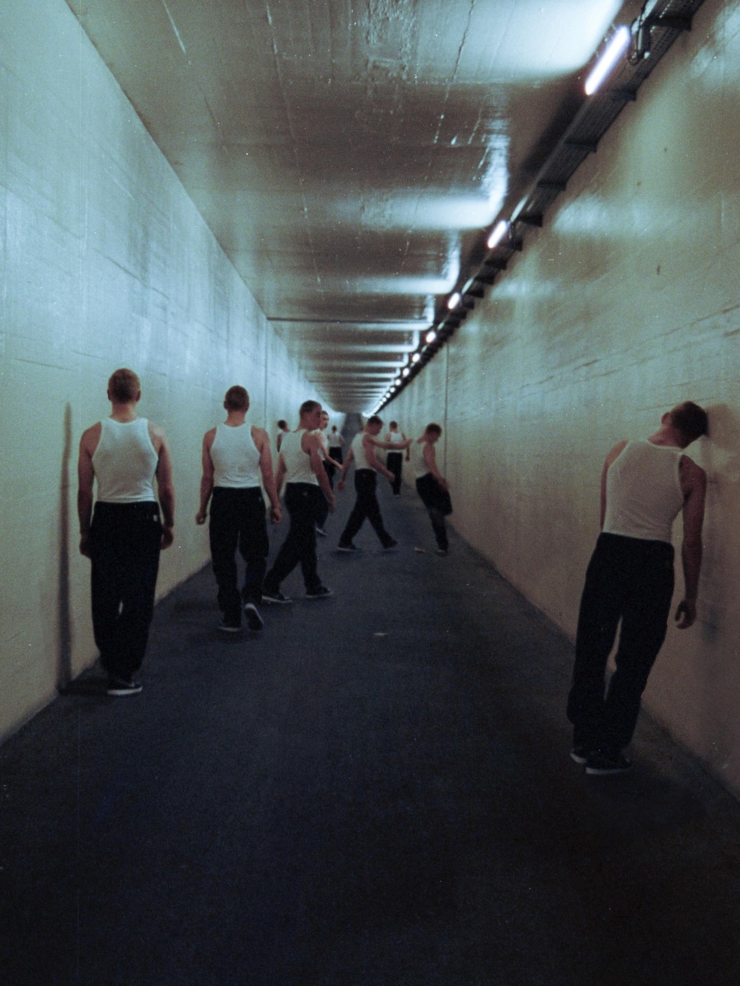 Several men in white tank tops and dark pants are walking and standing in a long, dimly lit tunnel.