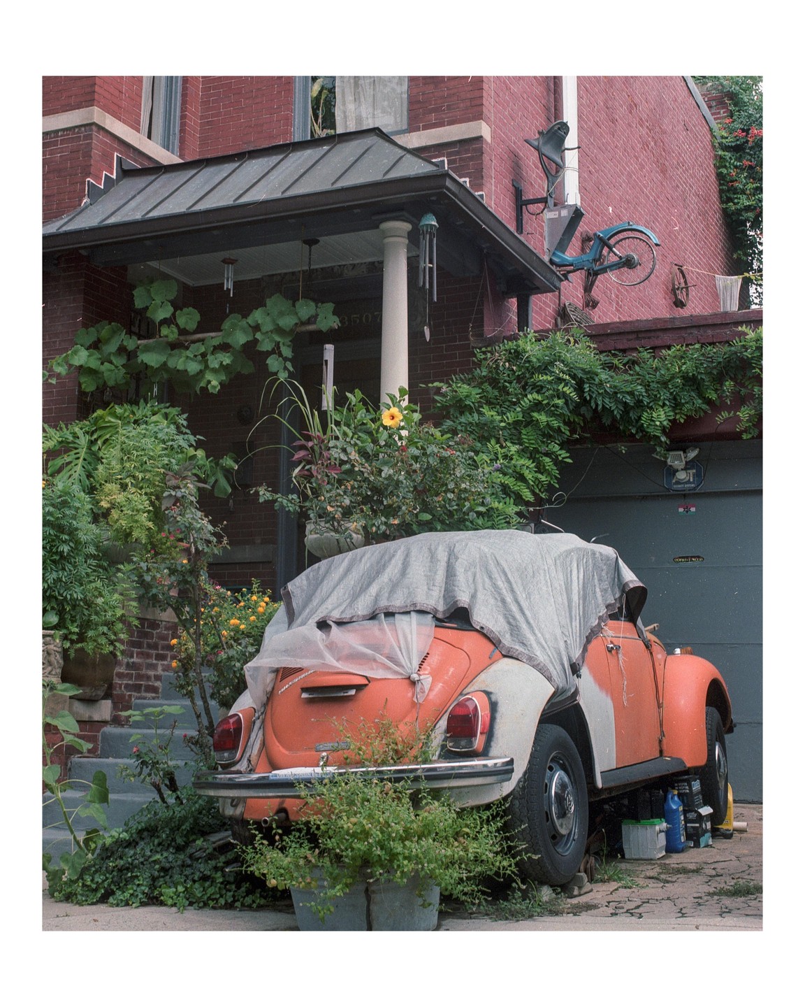 An orange car covered with a tarp is parked in front of a brick house with many plants.