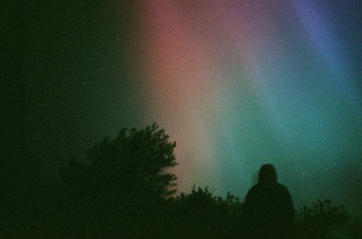 A person stands silhouetted against a colorful night sky with trees in the foreground.