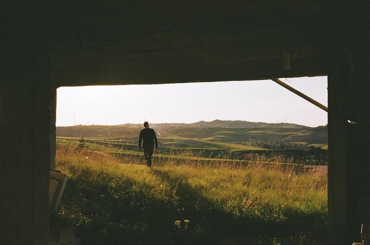 A man walks through a field of tall grass towards rolling hills under a bright sky, framed by a dark structure.