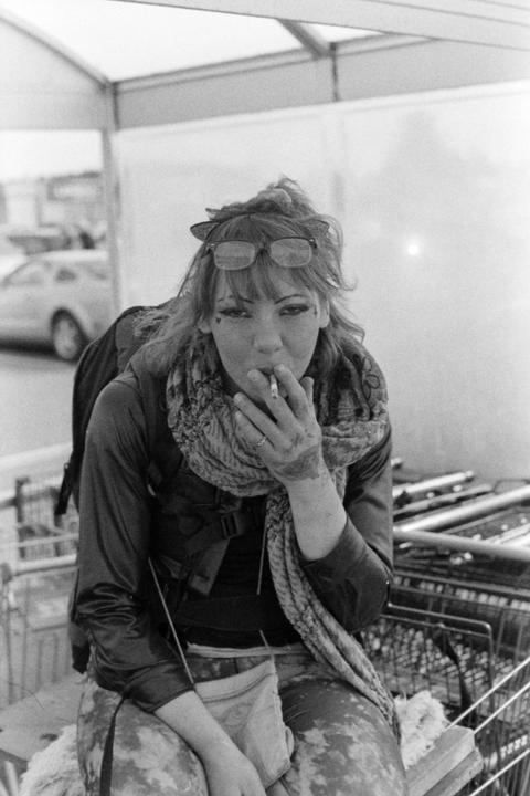 A woman with glasses on her head and a scarf around her neck sits and smokes a cigarette.