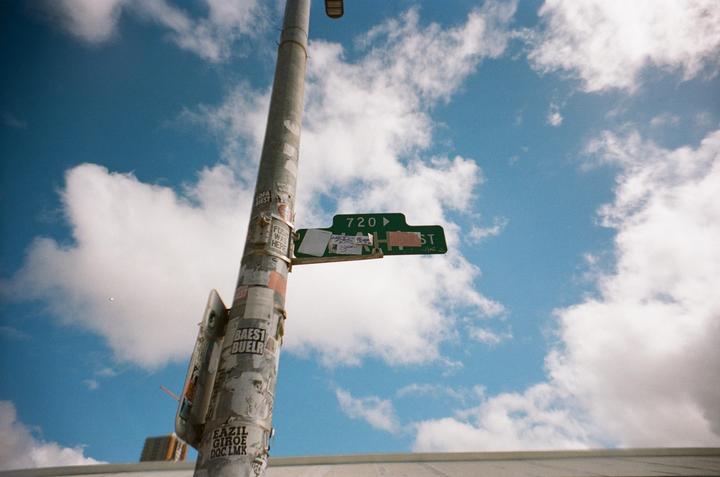 A street sign for 720 ST is attached to a utility pole covered in stickers and graffiti, set against a bright blue sky with white clouds.
