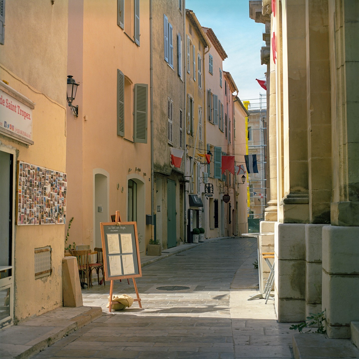 A narrow street in Saint Tropez is lined with buildings featuring shutters and flags, with a menu board and chairs visible on the left.