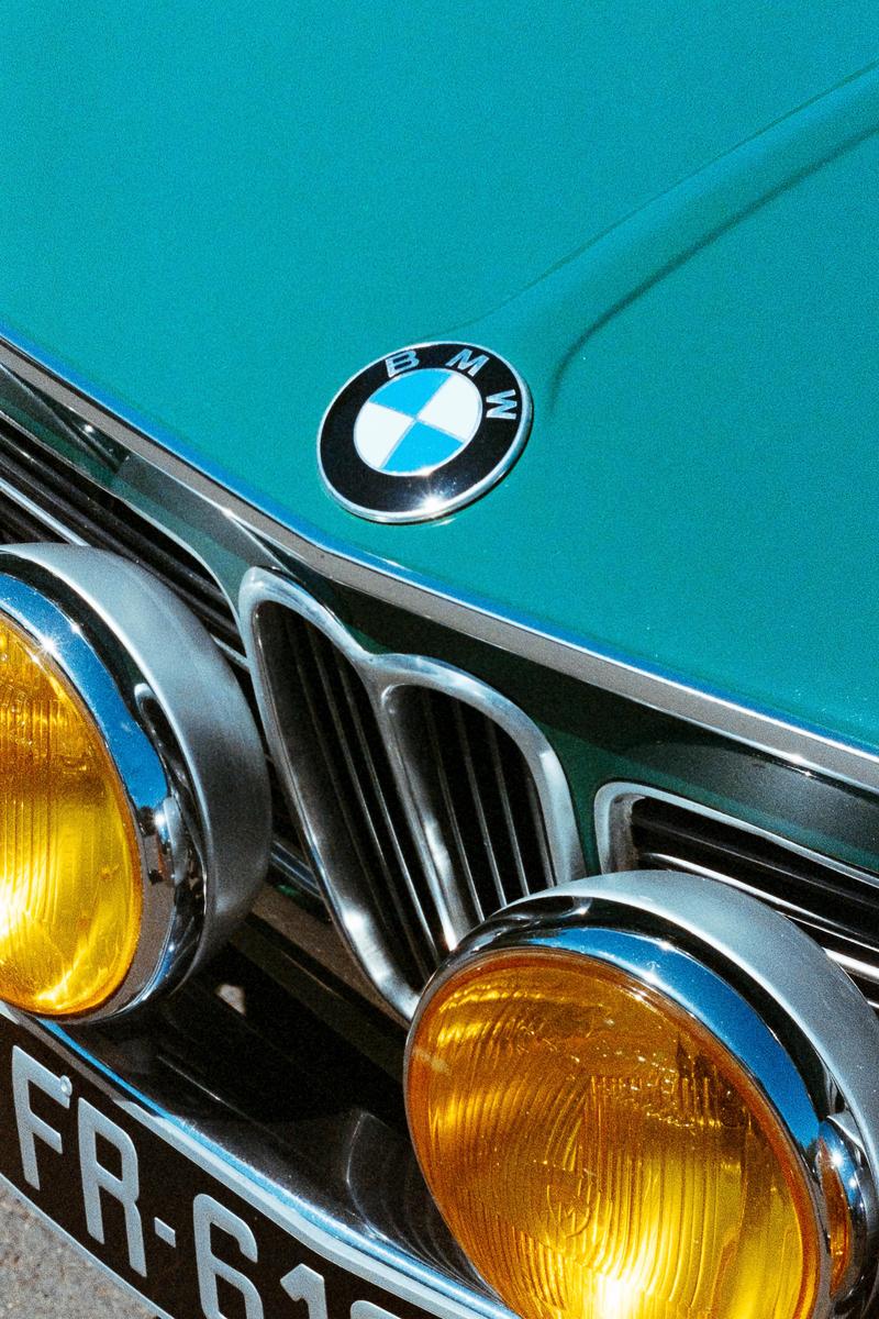 The front of a teal BMW car with yellow headlights and a visible license plate is shown.