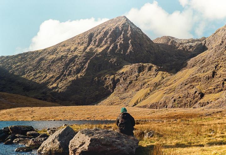 A person sits on a rock by a lake, looking at a large mountain under a blue sky with clouds.