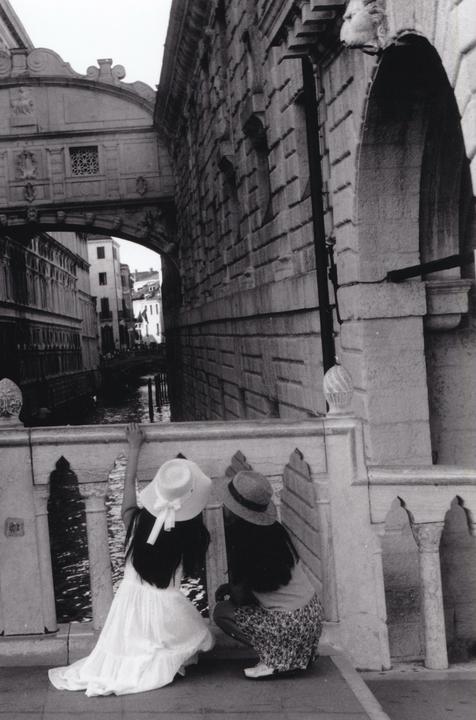 Two women wearing hats look over a stone bridge at a canal in Venice.