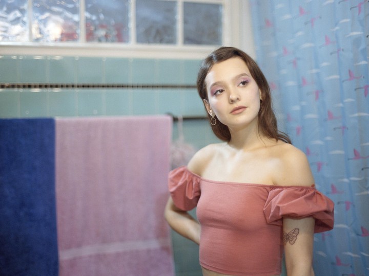 A young woman with pink eyeshadow and a butterfly tattoo on her arm looks to the right in a bathroom with blue tiles and a flamingo shower curtain.