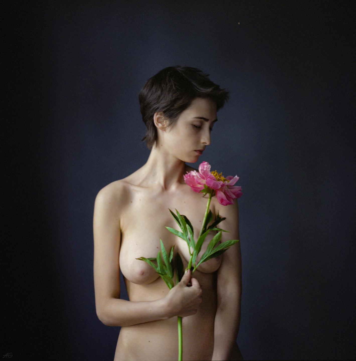 A woman with short hair holds a pink peony against a dark background.