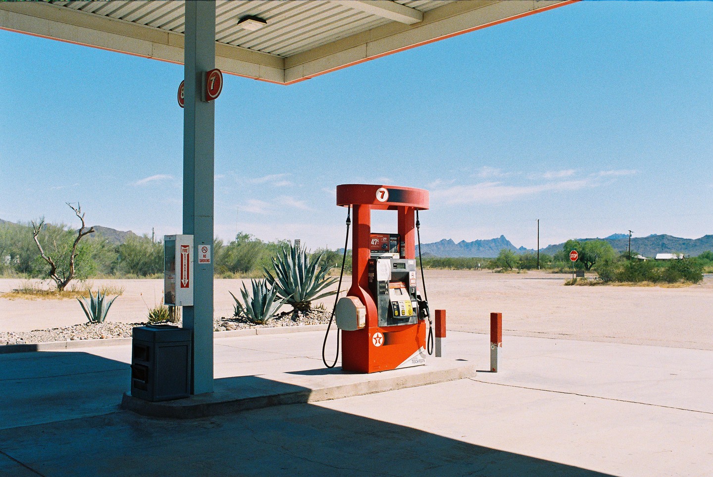 A red gas pump stands under a canopy at a gas station in a desert landscape with mountains in the distance.