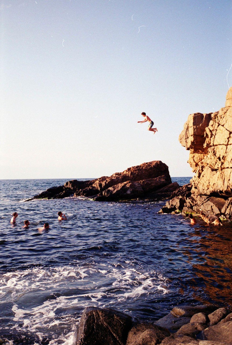 A man jumps from a cliff into the sea where other people are swimming.