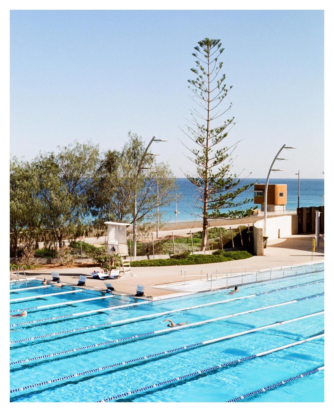 A swimming pool with several lanes is next to a beach with a tall, thin tree in the middle ground.