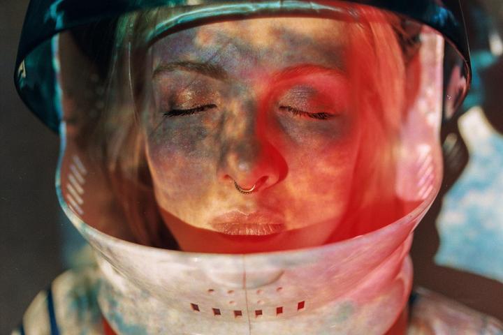 A woman with closed eyes wears a space helmet with colorful light projected onto her face.