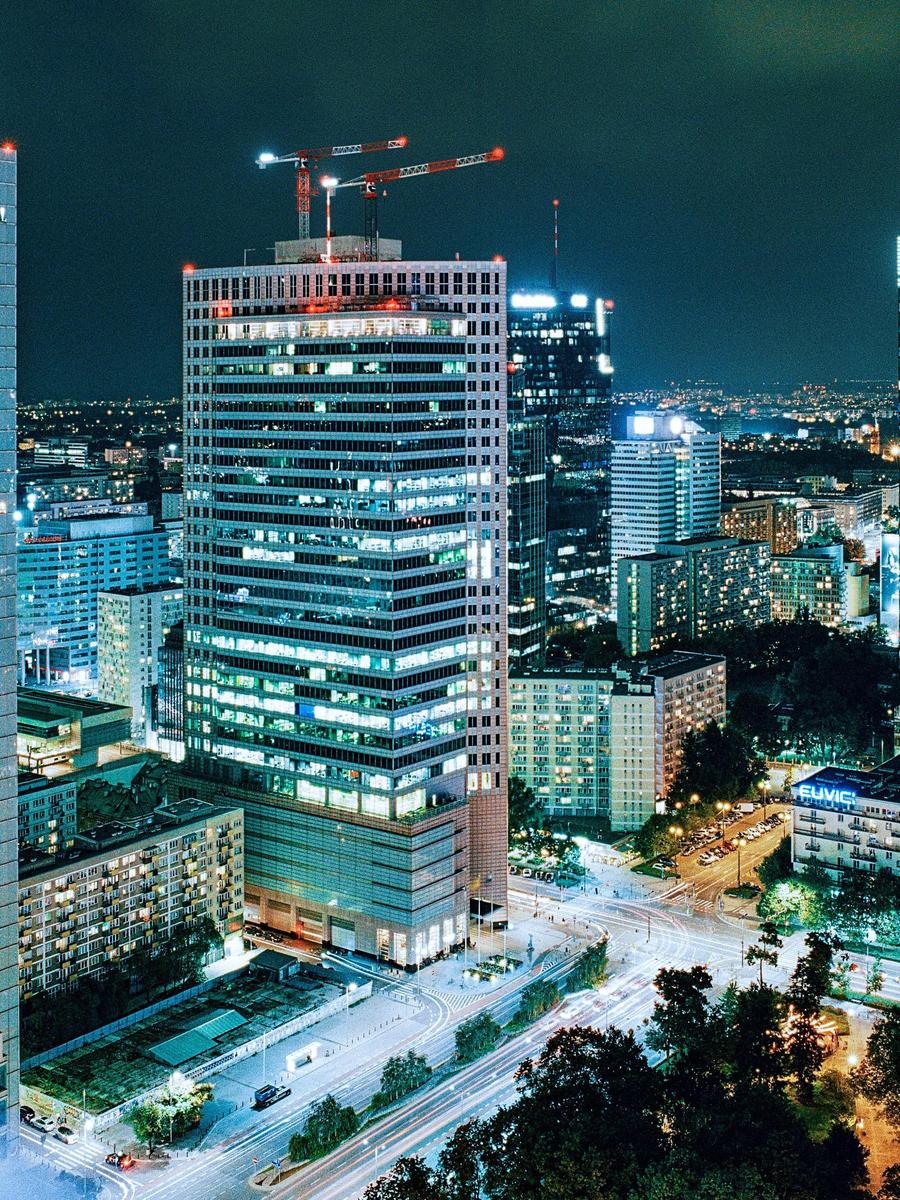 A tall building with construction cranes on its roof stands illuminated at night, surrounded by other city buildings and streets with light trails from vehicles.