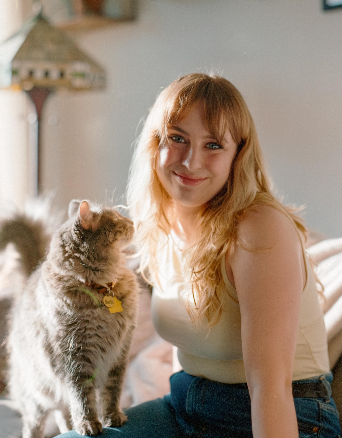 A woman with long blonde hair smiles at the camera while a cat stands on her lap.
