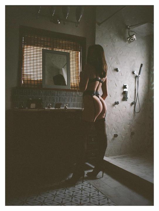A woman in lingerie stands with her back to the camera in a bathroom with a window and a shower.