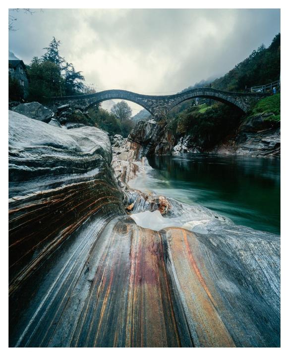 A stone bridge arches over a river with colorful rock formations in the foreground under a cloudy sky.