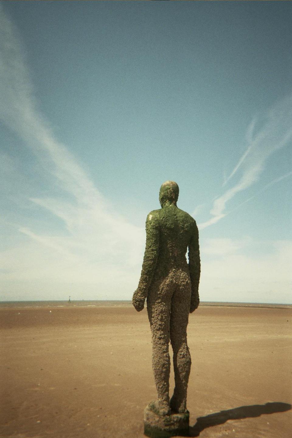 A statue of a man stands on a sandy beach looking out at the ocean under a blue sky with wispy clouds.