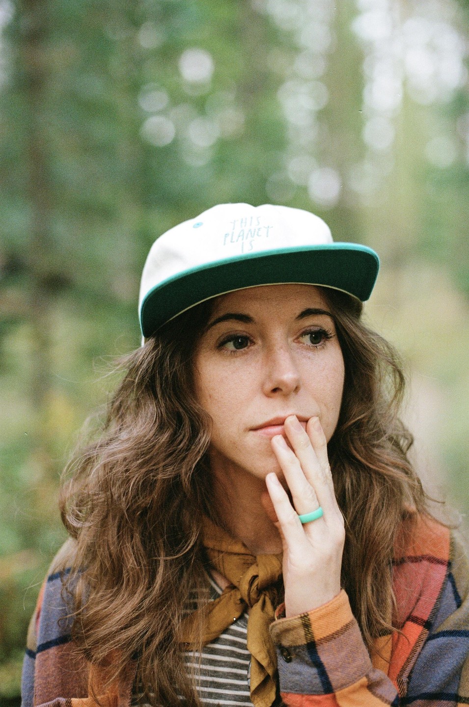 A woman with long wavy hair and a cap looks to the side, with her hand near her mouth.