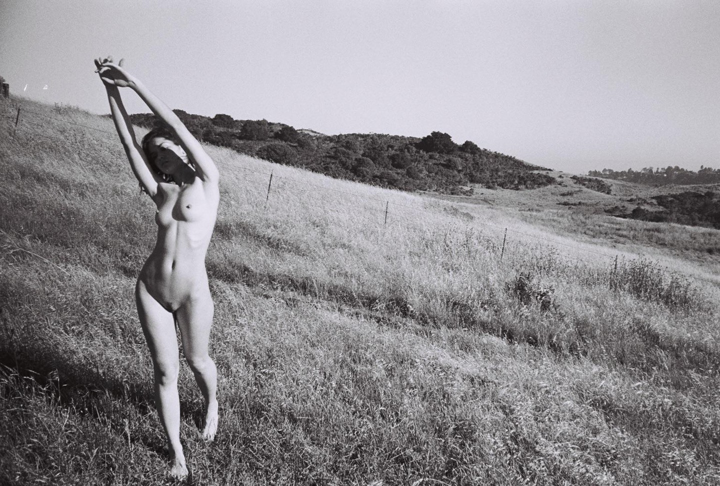 A nude woman stands in a field with her arms raised above her head.