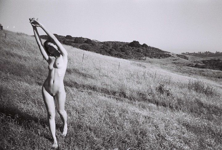 A nude woman stands in a field with her arms raised above her head.