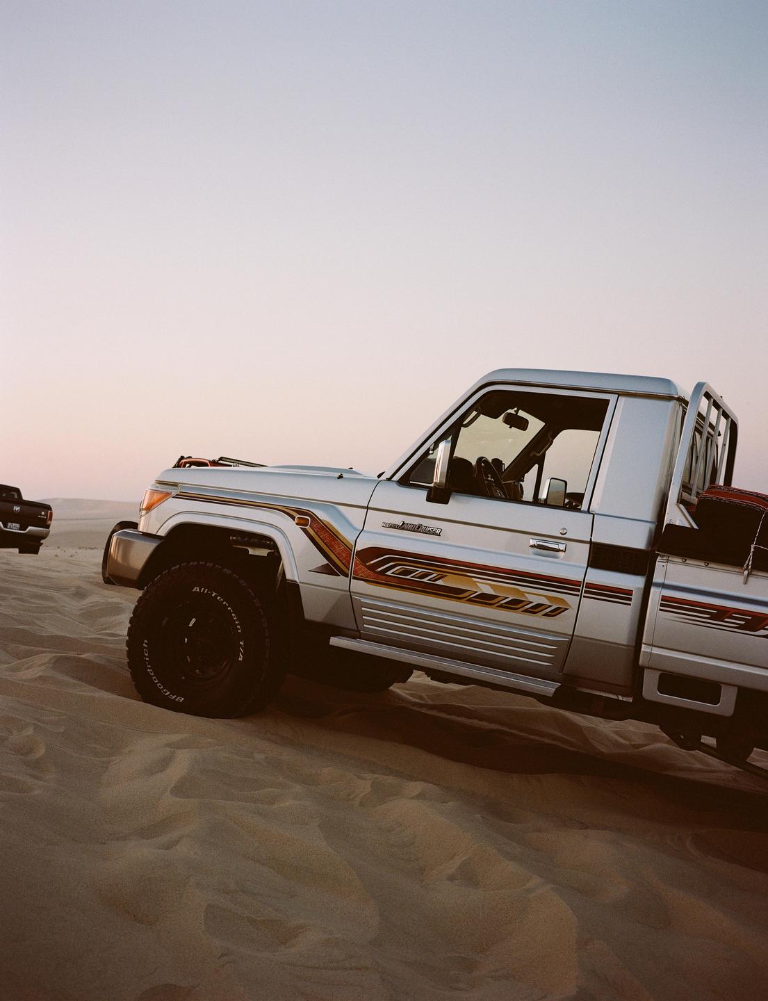 A silver pickup truck with striped decals is parked on sand dunes under a clear sky.