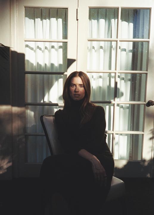 A woman sits in a chair in front of a glass door with light and shadow playing across her face and the door.