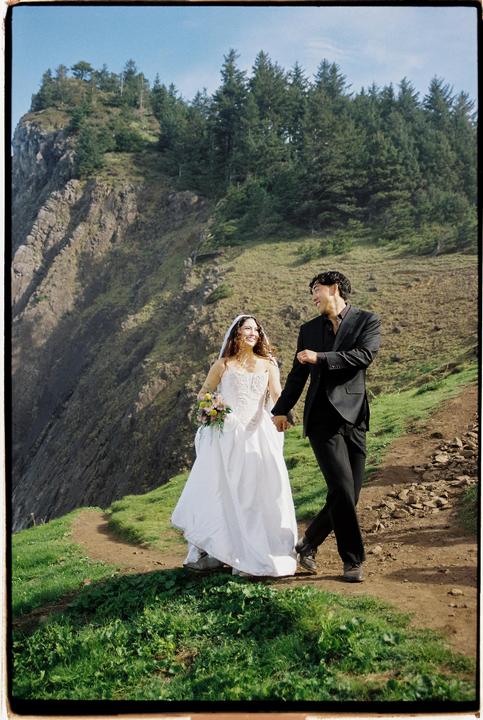 A couple walks hand-in-hand on a dirt path along a cliffside with a forest in the background.