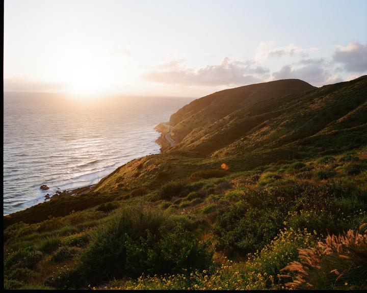 The sun sets over the ocean next to a winding road along a green hillside.