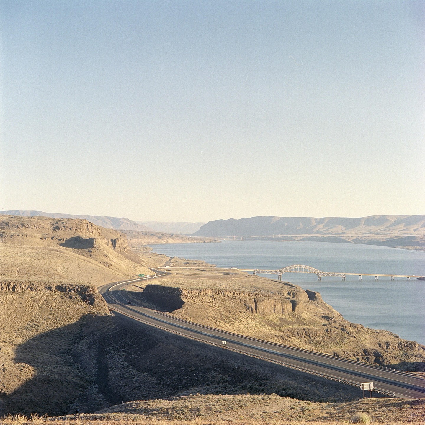 A highway curves along a dry, hilly landscape next to a large body of water with a bridge spanning it.