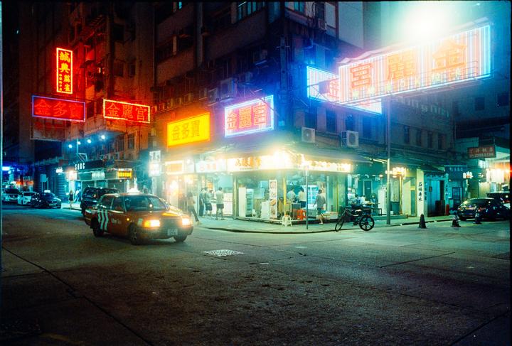 A street corner at night is illuminated by neon signs and storefront lights, with a taxi driving past.