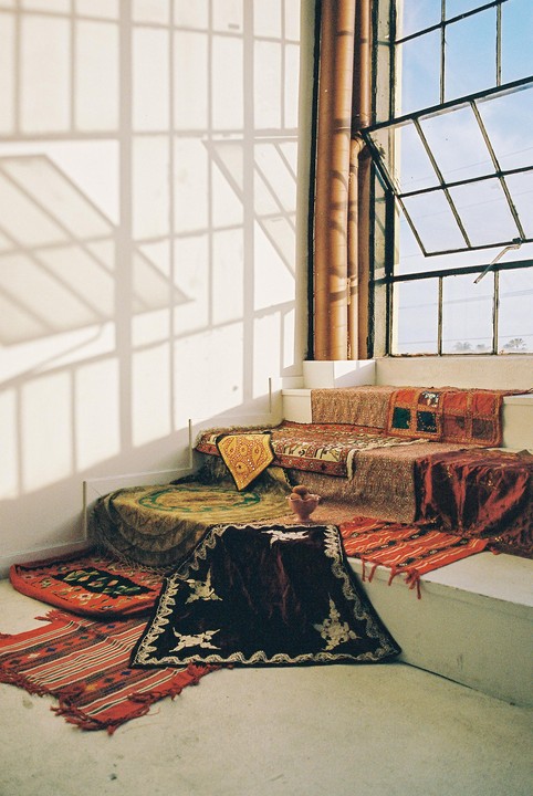 A room with a window and a wall with shadows from the window, with several rugs and pillows on the floor and on a raised platform.