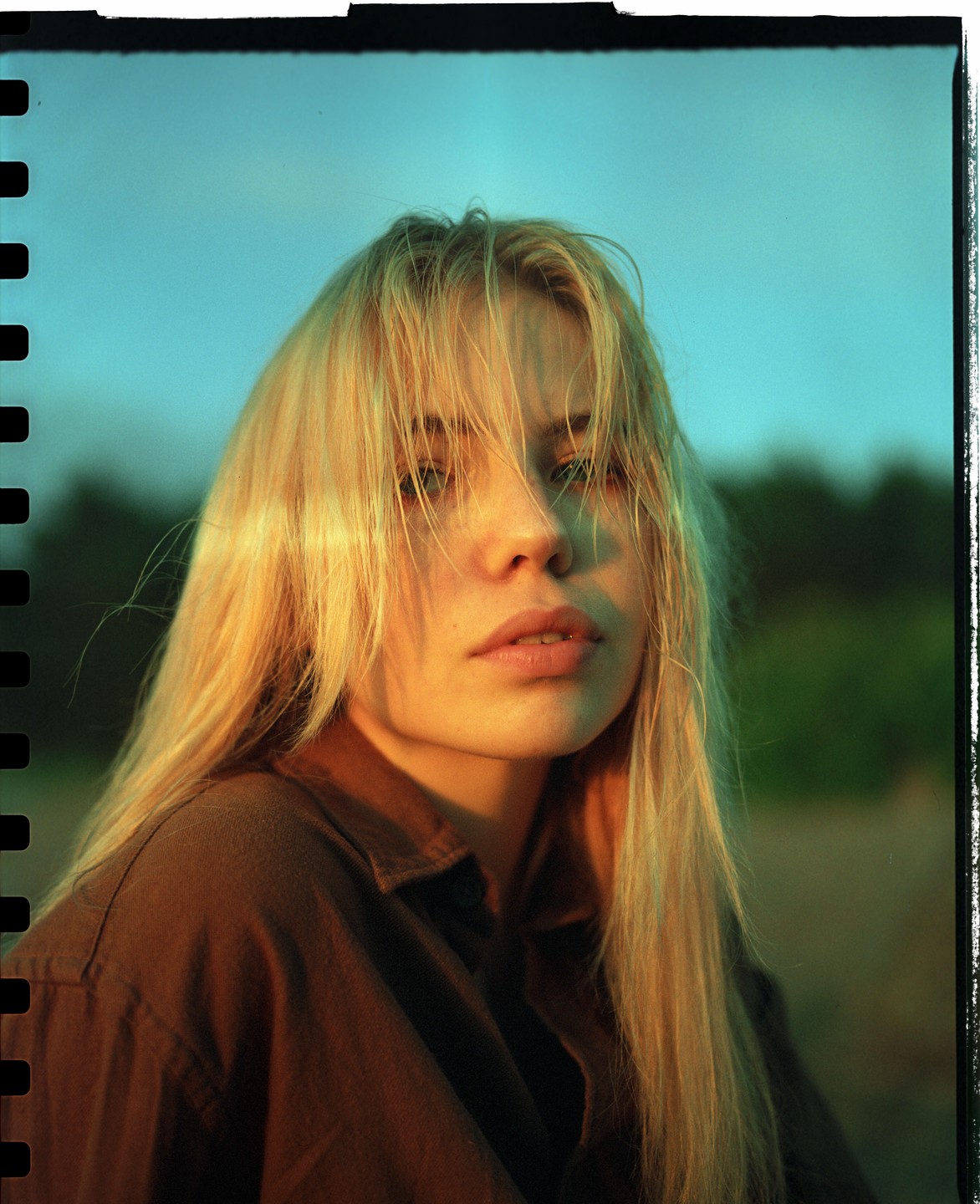 A woman with long blonde hair and a brown shirt looks directly at the viewer.