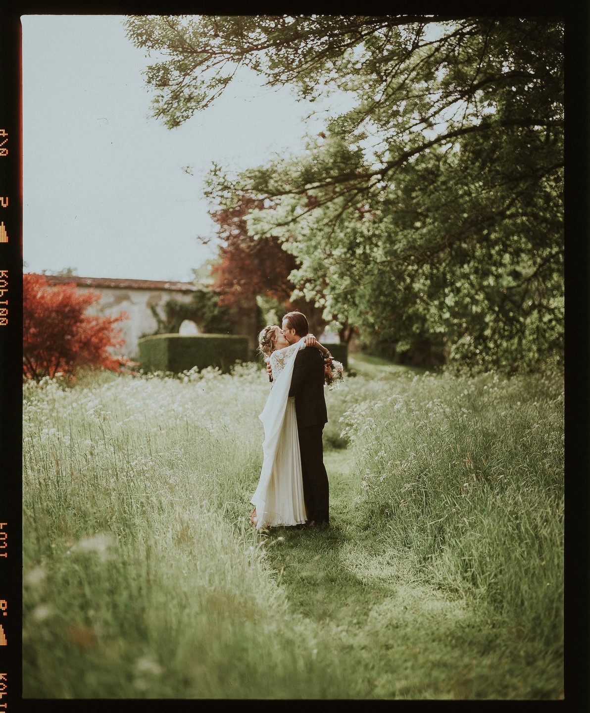 A couple embraces and kisses in a field of tall grass and wildflowers.