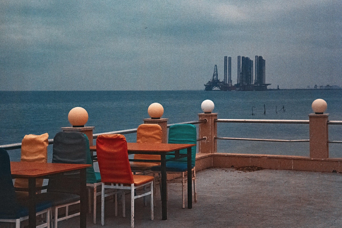 A cafe terrace with colorful chairs and tables overlooks the sea with an oil rig in the distance under a cloudy sky.