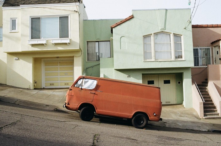 An orange van is parked on a steep street in front of three houses.