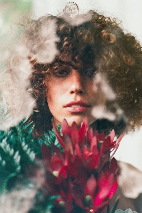 A woman with curly hair is visible through a double exposure with red and green foliage.