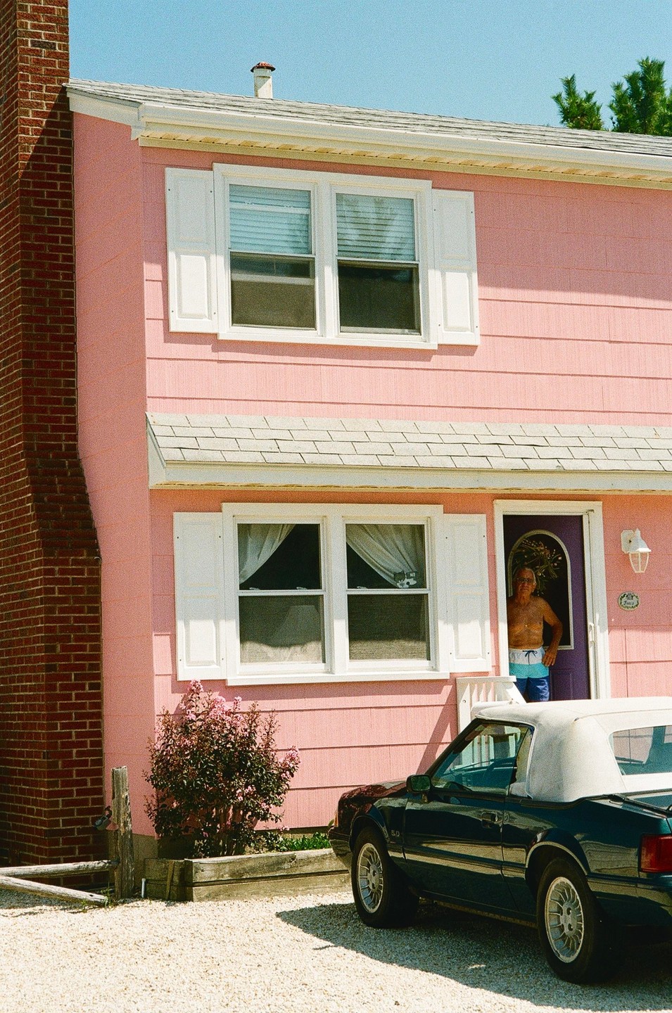 A man stands in the doorway of a pink house with a dark green convertible parked in front.