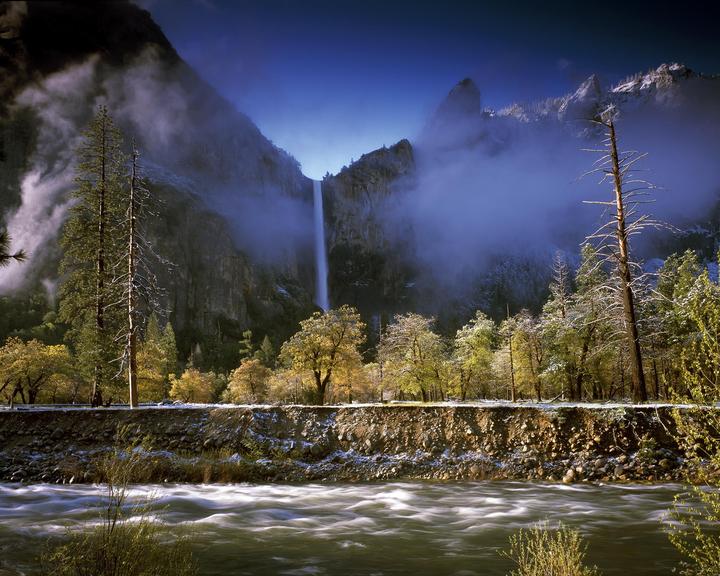 A waterfall cascades down a mountain into a river, with trees and a dusting of snow on the ground.