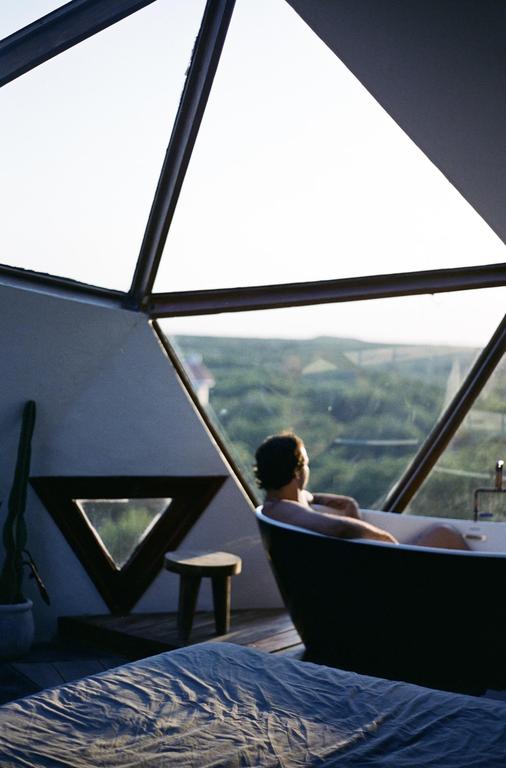 A man sits in a bathtub looking out a triangular window at a green landscape.