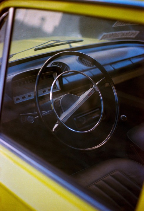 The interior of a yellow car is visible through the windshield and side window, showing the steering wheel, dashboard, and driver's seat.