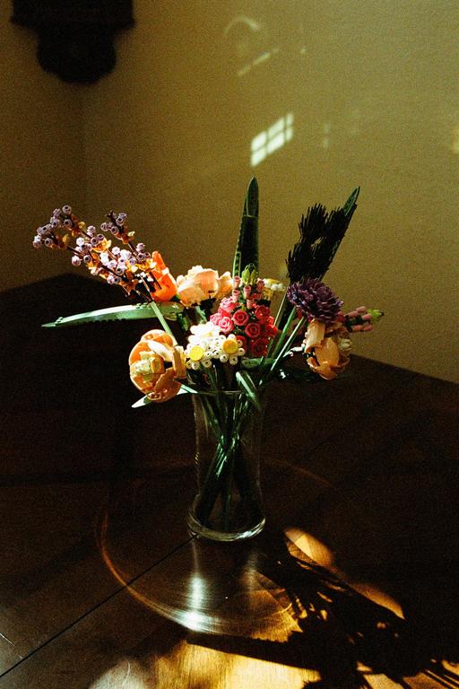 A bouquet of colorful lego flowers sits in a glass vase on a wooden table, illuminated by a window's light.