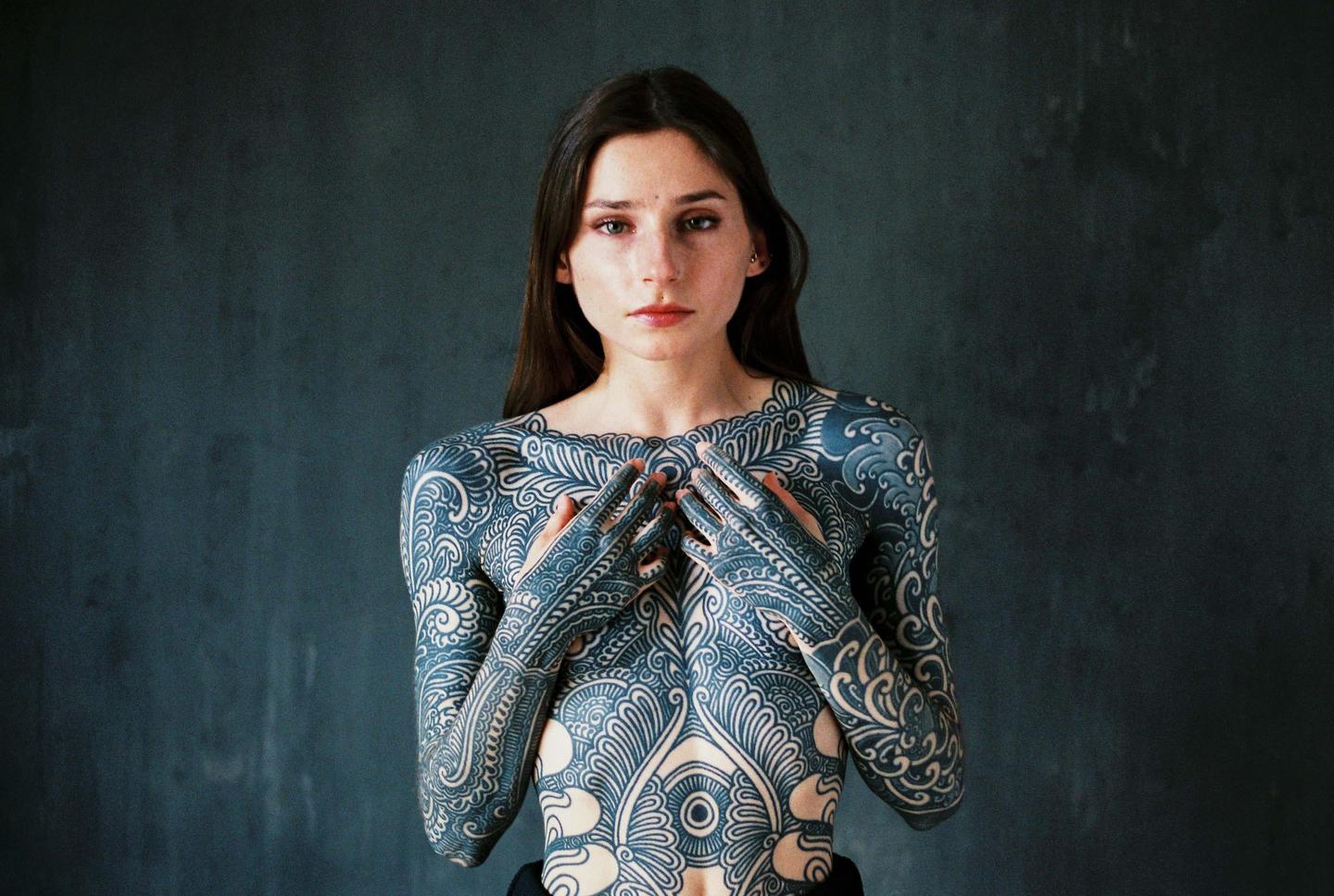 A woman with intricate blue body art covering her torso and arms looks directly at the viewer.