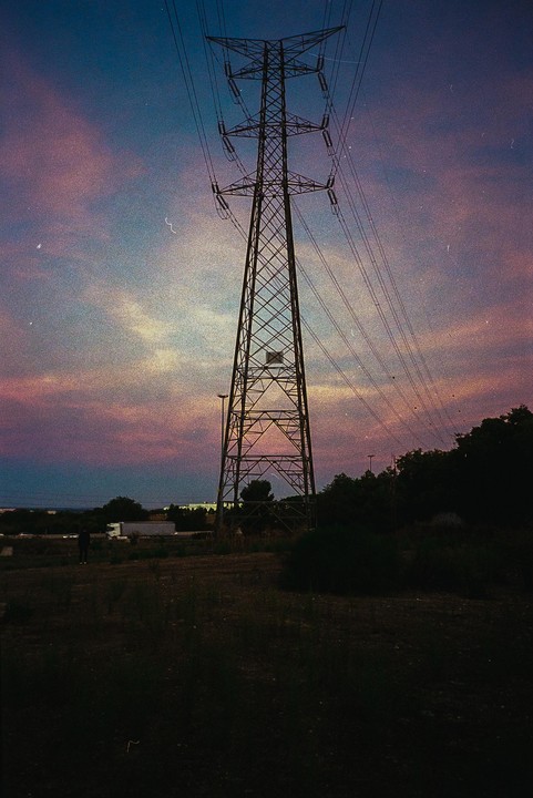 A tall electricity pylon stands against a colorful sky at dusk, with a person in the distance.