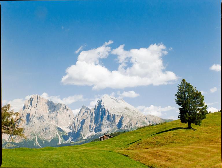 A small wooden hut sits on a green hillside with a single tree, overlooking a mountain range under a blue sky with clouds.