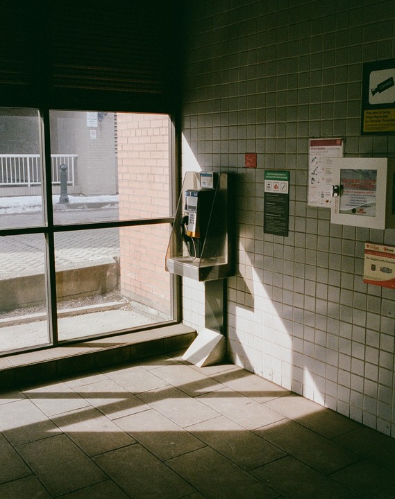 Sunlight streams through a large window onto a tiled floor and wall next to a payphone inside Dufferin Station.